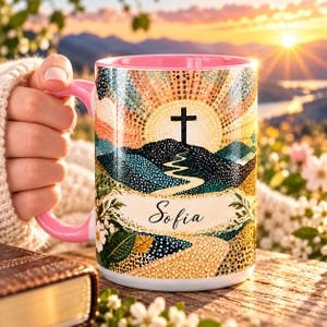 May include: A ceramic mug with a pink handle and interior, featuring a colorful graphic of a sunrise over rolling hills, a black cross, and a winding path. The name Sofia is printed in black script on a light banner across the center.