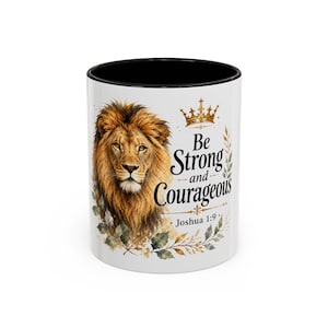 May include: A white ceramic mug with a black interior and rim. The exterior features a detailed illustration of a lion's head, a gold crown, and green foliage. The text reads: Be Strong and Courageous Joshua 1:9 in black, stylized lettering.