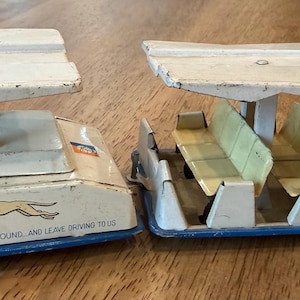 Könnte beinhalten: Zwei Vintage-Spielzeugbusse aus Metall mit weißen Dächern und blauen Unterteilen. Eine Einheit zeigt den Text Glide-a-ride und Go Greyhound, and leave driving to us mit einer Grafik eines laufenden Hundes. Die andere Einheit hat Reihen hellgelber Sitze.