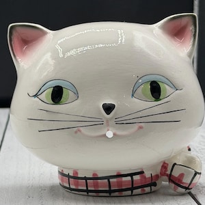 Puede incluir: Una figura de cerámica blanca con forma de cabeza de gato, con ojos verdes y sombra de ojos azul, nariz pequeña negra y bigotes negros. El gato tiene el interior de las orejas y la boca de color rosa. Una banda de cuadros rojos y blancos rodea el cuello.