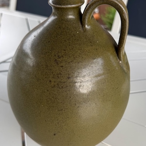May include: A round, olive green ceramic jug with a smooth, speckled glaze finish. The vessel features a narrow, flared neck opening and a single curved handle attached to the side, extending from the shoulder to the upper neck area.