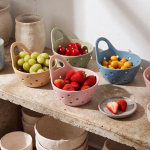 Puede incluir: Un conjunto de cuatro cuencos de cerámica para bayas en tonos pastel rosa, verde salvia, azul polvo y beige, cada uno con un asa lateral y orificios de drenaje. Los cuencos contienen fresas frescas, uvas verdes, tomates cherry y uchuvas.