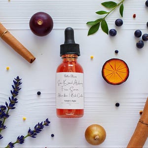 May include: A clear glass bottle with a black dropper, filled with a reddish liquid, is centered on a white wooden surface. The label reads "Sun Kissed Alchemy Face Serum". Cinnamon sticks, berries, and a sliced orange are arranged around the bottle.