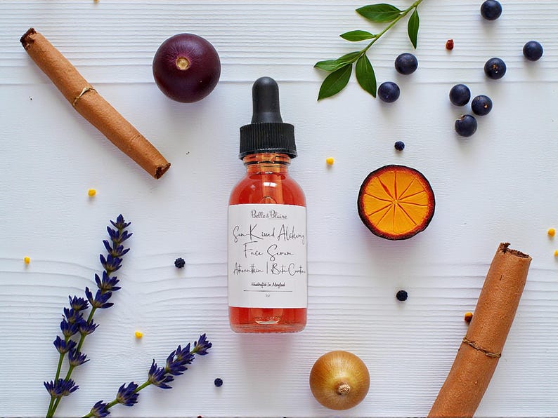 May include: A clear glass bottle with a black dropper, filled with a reddish liquid, is centered on a white wooden surface. The label reads "Sun Kissed Alchemy Face Serum". Cinnamon sticks, berries, and a sliced orange are arranged around the bottle.