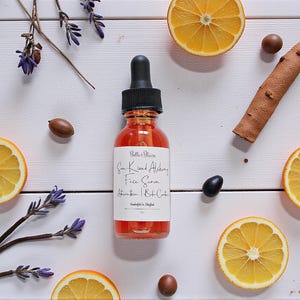 May include: A glass bottle of face serum with a black dropper, surrounded by orange slices, lavender sprigs, and spices on a white wooden surface. The serum has a red hue, and the label reads "Sun Kissed Alchemy Face Serum".