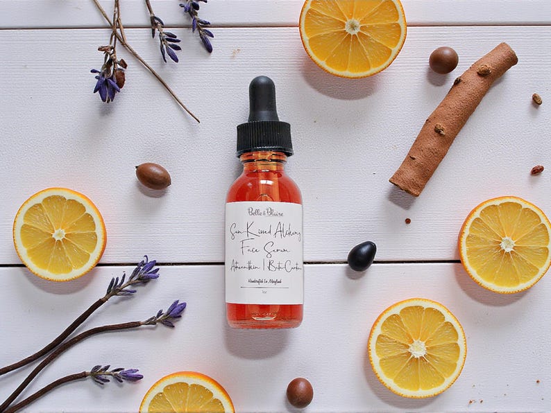 May include: A glass bottle of face serum with a black dropper, surrounded by orange slices, lavender sprigs, and spices on a white wooden surface. The serum has a red hue, and the label reads "Sun Kissed Alchemy Face Serum".