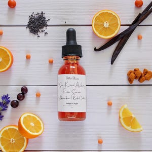 May include: A bottle of face serum with a black dropper, surrounded by orange slices, vanilla beans, and other ingredients on a white wooden surface. The label reads "Sun Kissed Alchemy Face Serum".