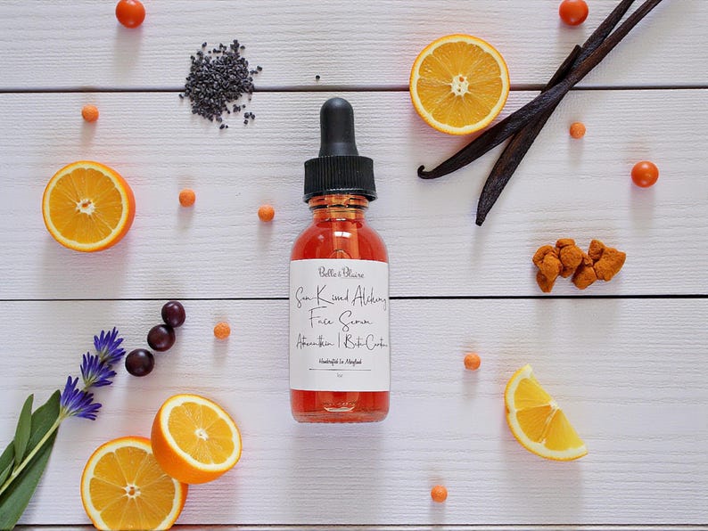 May include: A bottle of face serum with a black dropper, surrounded by orange slices, vanilla beans, and other ingredients on a white wooden surface. The label reads "Sun Kissed Alchemy Face Serum".
