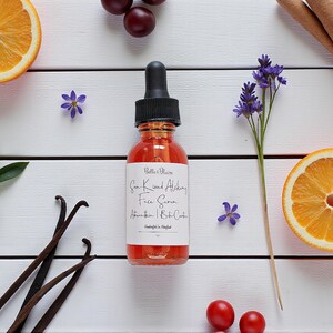 May include: A bottle of Sun Kissed Alchemy Face Serum with a black dropper, surrounded by orange slices, vanilla beans, cinnamon sticks, and purple flowers on a white wooden surface. The label reads "Belle & Blaire".
