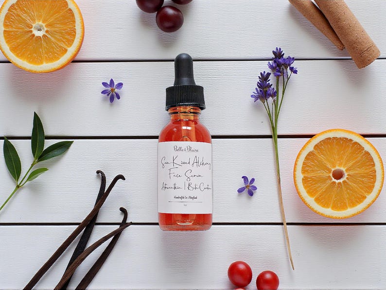 May include: A bottle of Sun Kissed Alchemy Face Serum with a black dropper, surrounded by orange slices, vanilla beans, cinnamon sticks, and purple flowers on a white wooden surface. The label reads "Belle & Blaire".