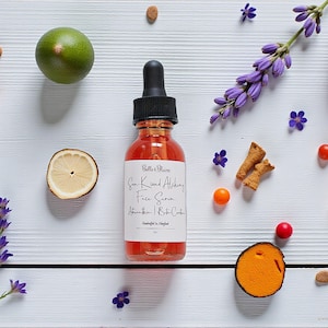 May include: A clear glass bottle with a black dropper top, filled with a reddish liquid, sits on a white wooden surface. The label reads "Sun Kissed Alchemy Face Serum". The image is decorated with lavender, citrus, and small orange berries.