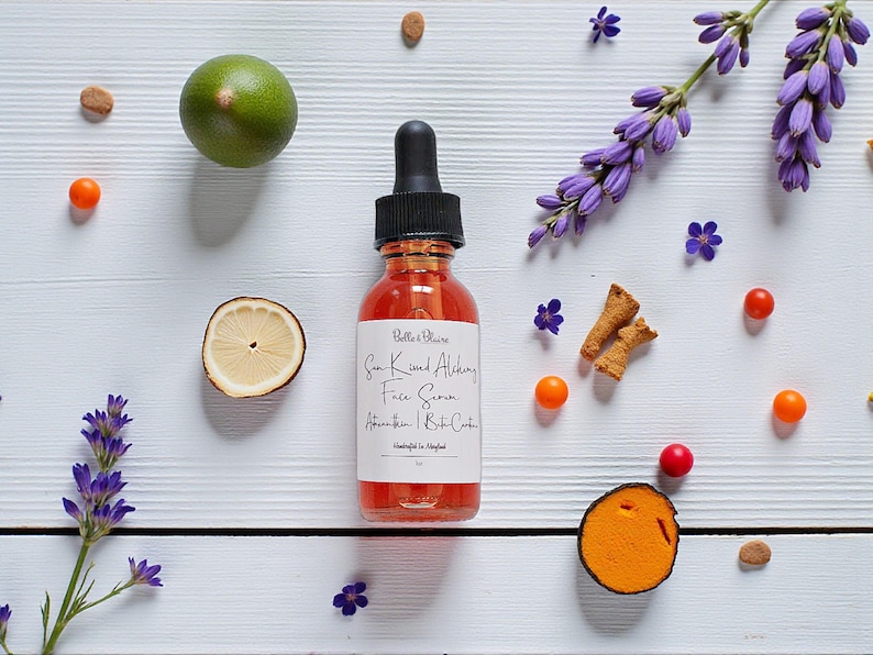 May include: A clear glass bottle with a black dropper top, filled with a reddish liquid, sits on a white wooden surface. The label reads "Sun Kissed Alchemy Face Serum". The image is decorated with lavender, citrus, and small orange berries.