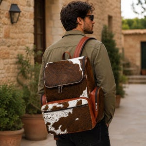 Könnte beinhalten: Ein Rucksack mit braun-weißem Kuhfellmuster und strukturierter Felloberfläche. Die Tasche verfügt über ein großes Hauptfach mit Klappverschluss, eine kleinere Reißverschlusstasche auf der Vorderseite und braune Lederseitenteile mit silbernen Reißverschlüssen.