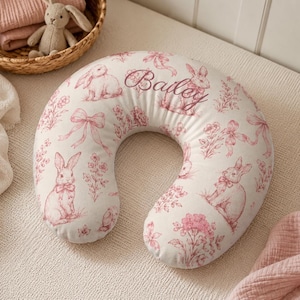 Puede incluir: Una almohada de lactancia en forma de C con una base de tela blanca y un patrón repetitivo de toile rosa con conejos, lazos y flores. El nombre Bailey está bordado en una caligrafía cursiva de color rosa en la parte superior central.