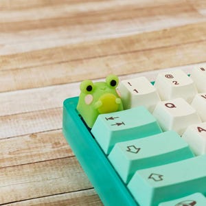 May include: A green and white keyboard with a frog-shaped keycap. The keycap is light green with black eyes and a yellow belly. The keyboard keys are white and light green.