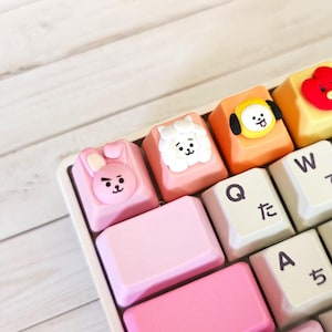 May include: A mechanical keyboard featuring custom artisan keycaps with sculpted 3D character faces including a pink rabbit, a white sheep, a yellow dog, and a red heart shape, alongside standard white keys with black English and Japanese characters.