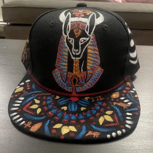 Pode incluir: Um boné de beisebol preto com um gráfico bordado detalhado da divindade egípcia Anúbis no painel frontal. A aba reta é decorada com um padrão colorido e intrincado em azul, vermelho, dourado e branco, incorporando vários símbolos.