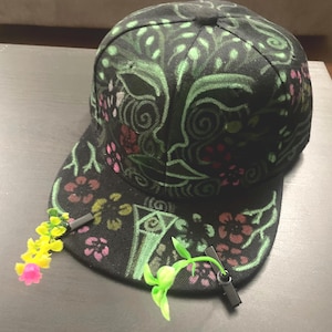 Puede incluir: Una gorra de béisbol negra con un diseño pintado a mano de líneas verdes en espiral y pequeñas flores rojas y rosas. Dos pequeños clips de plástico están sujetos a la visera, cada uno sosteniendo un tallo de flor artificial con pétalos amarillos y rosas.