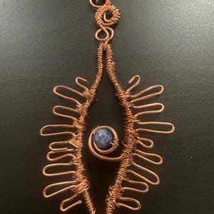 May include: A pendant crafted from copper wire shaped into an elongated eye-like frame with multiple radiating loops. A single round, blue and white mottled stone is secured within a spiral of copper wire at the center of the frame.