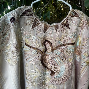 May include: A beige, striped tunic featuring intricate embroidery of a central bird with outstretched wings, surrounded by floral and geometric patterns in shades of brown, gold, and light blue, hanging on a white plastic clothes hanger.