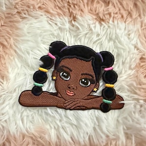 May include: An embroidered patch featuring a dark-skinned character with black hair styled in two high buns with colorful hair ties. The character has large brown eyes, long eyelashes, peach-colored lips, and small yellow star-shaped earrings.