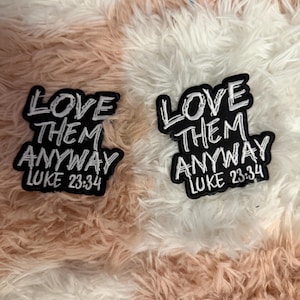 May include: Two black embroidered patches with white stitched text that reads LOVE THEM ANYWAY LUKE 23:34. The patches feature a bold, block-style font with a white border, showcasing a clear, high-contrast design for easy readability.