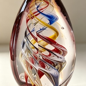 May include: A clear glass paperweight in an egg shape featuring an internal swirling design of red, yellow, blue, and white glass ribbons. The solid glass object contains small air bubbles throughout the structure and has a smooth, polished surface.