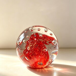 Puede incluir: Un pisapapeles esférico de vidrio transparente que presenta un núcleo rojo vibrante con remolinos y burbujas de aire suspendidas en su interior. La esfera de vidrio tiene una superficie lisa y pulida que refleja la luz, mostrando el color rojo intenso.
