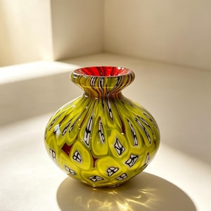 May include: A small, rounded glass vase featuring a vibrant chartreuse yellow body with a repeating pattern of white and black teardrop-shaped inclusions. The interior rim of the vase is a solid, deep red, contrasting with the yellow exterior.