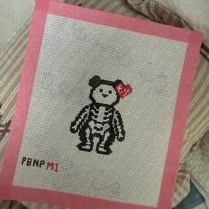 May include: A needlepoint canvas featuring a black and white bear with a visible skeleton design and a red heart on its left ear. The canvas has a pink border and the text PBNP M1 printed in black and red in the bottom left corner.