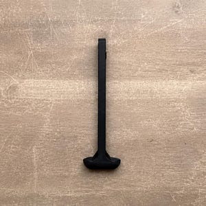 May include: A black T-shaped handle with a long, slender vertical shaft and a wider, rounded horizontal grip at the base. The object features a matte finish and a solid, uniform construction, presenting a simple, functional design for manual operation.