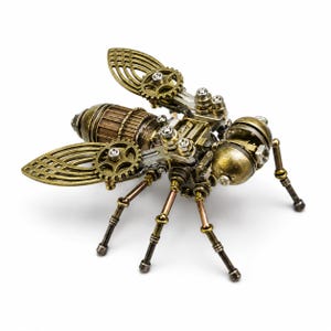 Può includere: Una scultura di insetto meccanico realizzata con componenti in metallo color ottone e argento. La figura presenta ali a forma di ingranaggio, un corpo cilindrico segmentato, una testa arrotondata e sei zampe sottili articolate in metallo.