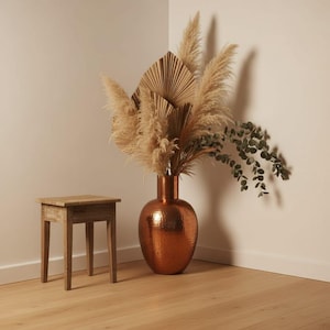 May include: A large, hammered copper vase with a rounded body and narrow neck, filled with dried pampas grass, beige fan-shaped palm leaves, and a sprig of green eucalyptus, positioned next to a small, light-colored wooden four-legged stool.