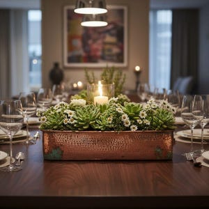 May include: A rectangular hammered copper planter box filled with green succulents and small white flowers, featuring a central glass candle holder with a lit white pillar candle, positioned as a centrepiece on a dark wood dining table.