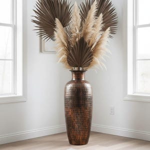 Könnte beinhalten: Eine hohe, bodenstehende Vase aus gehämmertem Kupfer mit dunklem, verwittertem Finish. Die Vase ist mit einem Arrangement aus getrockneten, fächerförmigen dunkelbraunen Palmblättern und hohen, federartigen beigen Pampasgraswedeln gefüllt.