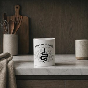 K&ouml;nnte beinhalten: Ein wei&szlig;er Keramikbecher mit einer schwarzen Grafik einer gewundenen Schlange, umgeben von floralen Linienzeichnungen. Der Text "I don't trust nobody (cc'd your boss)" ist in einer schwarzen gotischen Schrift &uuml;ber der Schlange aufgedruckt.