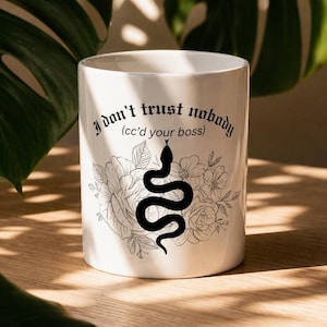 K&ouml;nnte beinhalten: Ein wei&szlig;er Keramikbecher mit einer schwarzen Grafik einer Schlange, die sich vor einer Blumenillustration windet. Der Text "I don't trust nobody (cc'd your boss)" ist in einer schwarzen gotischen Schrift &uuml;ber dem Schlangen- und Blumenmotiv gedruckt.