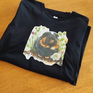May include: Black t-shirt featuring a white graphic of a guinea pig with brown and white fur surrounded by flowers and greenery.