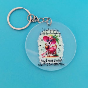May include: A circular, pale blue keyring featuring a flamingo design. The flamingo wears sunglasses and a hat, holding a drink, and sitting in a beach chair. Text reads, "I had a lot of stuff to do today. Now I have a lot of stuff to do tomorrow."