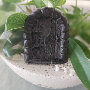 May include: A miniature fairy door, painted in dark grey, with a heart-shaped cutout. The door has a textured wood grain appearance and is framed by rounded stone-like elements. A small, decorative item for fairy gardens.