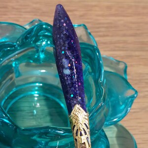 May include: A close-up of a purple and gold pen with glitter embedded in the purple resin. The pen is sitting in a turquoise blue glass rose.