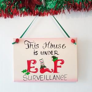 May include: A pink and cream wooden sign with the text "This House is Under ELF Surveillance" in black cursive writing. The letters "ELF" are in red and decorated with a green elf hat, a red boot, and a white and green striped stocking.