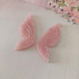 May include: Two pink glitter fairy wings, each with a delicate, detailed design. The wings are made of resin and have a shimmery finish.