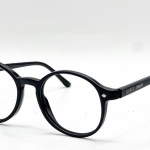 May include: Black round-frame eyeglasses with a glossy finish. The temples feature the text Giorgio Armani in white lettering. Small silver-toned diamond-shaped accents are visible on the front corners of the frame near the hinges.