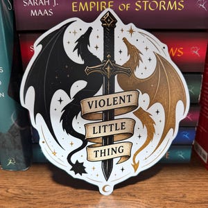 May include: A die-cut sticker featuring a central black sword flanked by a black dragon on the left and a gold dragon on the right. A beige ribbon banner wraps around the sword with the text VIOLENT LITTLE THING printed in black capital letters.