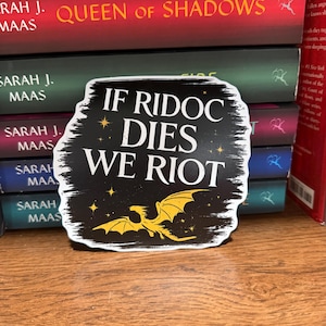 Könnte beinhalten: Ein schwarzer Aufkleber mit weißem Pinselstrich-Rand und dem Text IF RIDOC DIES WE RIOT in weißen Großbuchstaben. Das Design zeigt eine gelbe Drachensilhouette am unteren Rand sowie kleine gelbe Sternakzente auf dunklem Hintergrund.
