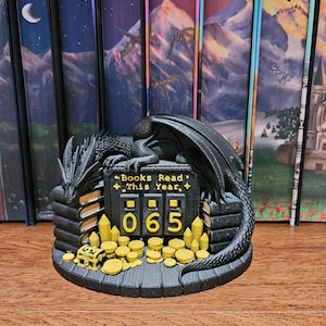 May include: A black dragon figurine resting on a circular base with stacks of books, gold coins, and crystals. A central black sign with yellow text reads Books Read This Year above a mechanical counter displaying the number 065 in yellow digits.