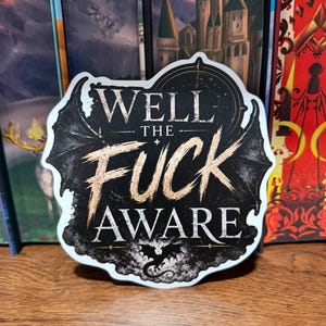 May include: A die-cut sticker featuring a black background with a white and gold-toned text design that reads WELL THE FUCK AWARE. The sticker includes graphic illustrations of bat-like wings on the sides and a small dragon silhouette at the bottom.