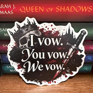 May include: A die-cut sticker featuring a skull wearing a crown, a dagger, and black roses against a dark, splattered background. The text reads, "I vow. You vow. We vow." in a white serif font, set against a backdrop of stacked book spines.