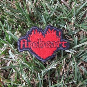 May include: An embroidered patch featuring a bright red heart shape with flame-like edges and a black border. The word fireheart is stitched across the center in a black, stylized lowercase font, creating a bold contrast against the red background.